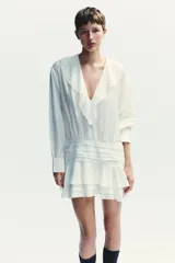 Vestido corto blanco de escote en V y manga larga con puños abotonados. Presenta volantes con bordados de encaje en el cuello, escote y falda. Cierre lateral con cremallera oculta.