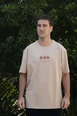 Remera color beige con cuello redondo y mangas cortas. Estampado frontal con tres hongos rojos y la palabra 'Tuya' debajo.