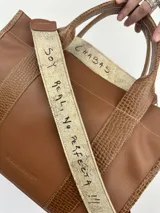Cartera marrón de cuero genuino con doble asa corta y correa bandolera estampada con la frase "Chabas, soy real, no perfecta".