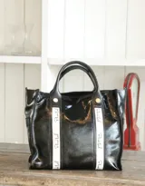 Cartera tipo bolso de cuero negro con acabado brillante, cuenta con doble asa de mano y detalles de cintas verticales en color crema con el nombre de la marca estampado. Incluye herrajes metálicos dorados.