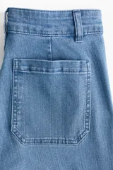 Jeans de tiro alto y corte acampanado, confeccionados en denim con leve elasticidad. Presentan cierre frontal con múltiples botones metálicos visibles, bolsillos de parche delanteros y diseño de perneras anchas desde la rodilla.