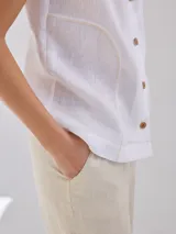 Camisa sin mangas de lino, color blanco, con cuello solapa, cierre frontal con botones y bolsillo pequeño en el pecho. Presenta costuras decorativas a la vista que definen la silueta.