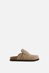Zueco color beige de símil gamuza con tira ajustable en el empeine y hebilla metálica plateada. Presenta plantilla de corcho y suela plana texturizada.