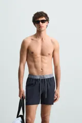 Bañador de hombre azul marino con cintura elástica gris y cordón ajustable. Presenta un pequeño texto estampado en relieve en la parte inferior izquierda.