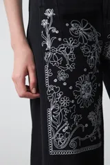 Pantalón de denim negro de corte recto y pierna amplia, con largo al tobillo. Presenta bordados florales en color blanco en la parte frontal superior y en la botamanga.
