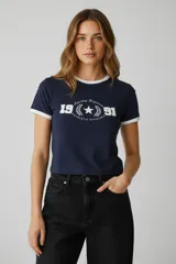 Remera de algodón con elastano, de corte holgado y manga corta. Presenta un diseño tipo ringer con cuello y bordes de mangas en color blanco contrastante sobre una base azul marino. Incluye un estampado gráfico frontal con el número 1991 y detalles de estilo deportivo.