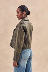 Campera corta color verde oliva de cuero sintético con cuello clásico, cierre frontal asimétrico con cremallera, bolsillos con cremallera y solapa, y detalles de costuras en los hombros.