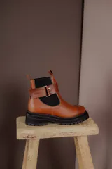 Bota tipo Chelsea de cuero marrón con suela track negra de goma. Presenta paneles elásticos laterales y una costura reforzada en la puntera. Incluye una correa de cuero desmontable con hebilla metálica.