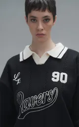 Sweater de punto negro con cuello polo en color crema, manga larga y diseño oversize. Presenta bordados en el frente con la inscripción 'Ravers', el número '90' y letras cursivas, con terminaciones en punto rib en puños y cuello.