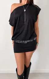 Vestido corto negro de estilo asimétrico con un hombro al descubierto, corte holgado en la parte superior y ajustado en la falda. Incluye un cinturón negro con ojales metálicos plateados.