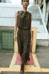 Vestido midi sin mangas de cuello redondo, con diseño de rayas verticales en tonos marrones. Presenta un detalle drapeado en el frente, volantes laterales y un bajo asimétrico. Cierre posterior con cremallera oculta.
