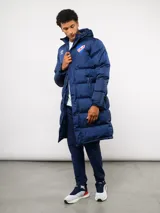 Parka larga acolchada color azul marino con capucha, cierre frontal con botones a presión y bolsillos laterales. Presenta el logo de Umbro y el escudo del Club Nacional de Football bordados en el pecho.