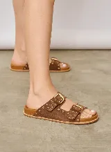 Sandalia chata de gamuza color marrón, con dos tiras cruzadas en el empeine, hebillas de metal dorado y detalles de ojales metálicos a lo largo de las tiras.