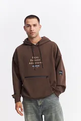 Canguro oversize color marrón, con capucha, cordones ajustables, bolsillo frontal con cierre y estampa frontal con texto y logo de la marca.