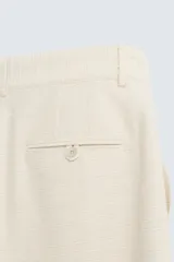 Bermuda de corte relajado color beige claro, confeccionada en tejido con textura. Presenta cierre frontal con cremallera y botón, bolsillos delanteros y bolsillos traseros de doble vivo con botón.