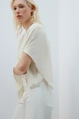 Camiseta tipo capa oversize de color blanco, confeccionada en hilatura de algodón 100%. Presenta cuello redondo, manga corta y un diseño de bajo asimétrico con caída pronunciada en uno de los laterales.
