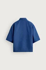 Camisa marrón de corte boxy fit, con tejido estructurado, cuello bowling, manga corta y un bolsillo aplicado en el pecho. Cuenta con cierre frontal de botones.