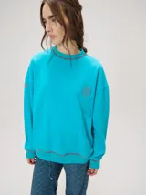 Buzo oversize color gris melange con corte crew, cuello redondo, mangas largas y detalles bordados en color verde en el cuello, puños y ruedo. Presenta el logo de la marca bordado en el pecho.