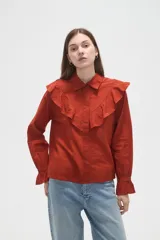 Camisa de algodón blanca con cuello camisero, cierre frontal de botones y volados bordados en el frente y hombros. Presenta mangas largas con puños acampanados y detalles bordados en el ruedo.