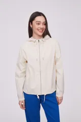 Campera liviana color crema con capucha, cierre frontal completo y cordones ajustables en el cuello y el ruedo. Cuenta con mangas largas con puños elásticos y bolsillos laterales.