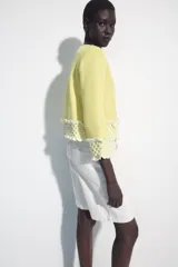 Chaqueta de punto color amarillo claro, con cuello redondo y mangas por debajo del codo. Presenta detalles de acabado en tejido crochet blanco en el ruedo y los puños, con cierre frontal de botones forrados al tono.