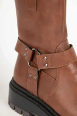 Bota estilo biker de cuero sintético color negro, con suela tipo plataforma dentada. Presenta una tira decorativa con argolla metálica y apliques de tachas en el tobillo.