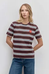 Remera de manga corta con cuello redondo, confeccionada en tejido de punto con diseño de rayas horizontales en tonos marrón, blanco y celeste. Presenta un pequeño parche circular rojo en el pecho.