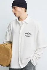 Camisa de corte relajado confeccionada en tejido fluido con mezcla de viscosa, color negro. Presenta cuello solapa, manga corta, cierre frontal con botones y un bordado a contraste en el pecho.