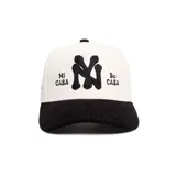 Gorro tipo cap de pana, color blanco con visera negra. Cuenta con el logotipo NY bordado en relieve en el centro y las frases "Mi Casa" y "Su Casa" bordadas a los lados.