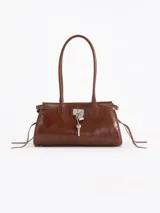 Cartera de hombro confeccionada en cuero vacuno marrón, con cierre superior y bolsillo interno. Presenta aplique metálico con candado y llavero colgante en el frente, además de tiras laterales decorativas.