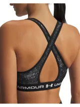 Top deportivo de soporte medio con diseño de espalda cruzada. Presenta un estampado jaspeado en tonos negros y grises, con una banda elástica inferior negra que incluye el logo de la marca en letras blancas. Confeccionado en tejido HeatGear de doble capa, de secado rápido y alta transpirabilidad.