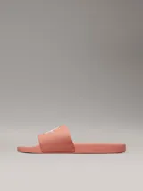 Ojotas tipo slide de color rosado, con base de EVA y correa ancha de lona de algodón reciclado. La correa presenta el logo de Calvin Klein Jeans bordado en blanco.