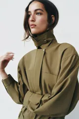 Vestido corto verde militar de cuello solapa y manga larga, con bolsillos de plastrón en el delantero y cierre frontal con cremallera oculta por sopata. Cuenta con un cinturón incorporado del mismo tejido con hebilla metálica y aberturas laterales en el bajo.