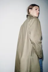 Trench coat de corte largo y diseño minimalista, confeccionado en tejido encerado color beige. Presenta cuello solapa, cierre frontal con botones oscuros y bolsillos laterales integrados.