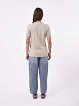 Remera de manga corta color beige claro con cuello redondo. Presenta un estampado gráfico central con un plato de pasta rodeado por un borde azul y blanco, sobre un fondo de franjas verticales rojas y blancas. El estampado incluye texto en cursiva en tonos turquesa y rojo.