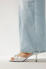 Sandalia de tacón fino color blanco, con tira delantera ancha con detalle de lazo y terminación en punta redonda.