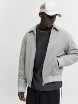 Campera bomber corta de paño gris claro, con cuello camisero, cierre frontal metálico completo y bolsillos laterales ribeteados. Presenta puños y cintura elasticados en rib acanalado.