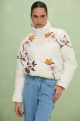 Campera puffer color blanco off-white con diseño acolchado, cuello alto y cierre frontal con botones a presión. Presenta bordados florales en tonos violeta, amarillo y rosa en la parte frontal y mangas.