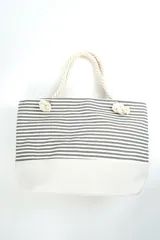 Bolso tipo tote de playa, con diseño de rayas horizontales en color gris y base blanca. Cuenta con asas de soga gruesa en color crudo con nudos decorativos.