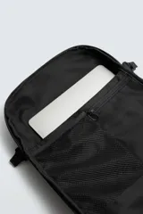 Mochila técnica de color negro con múltiples compartimentos, incluyendo dos bolsillos frontales con cierre, bolsillos laterales y compartimento principal acolchado para dispositivos electrónicos. Cuenta con parte posterior acolchada, banda de sujeción para equipaje, asa de mano y asas de hombro ajustables.