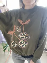 Buzo cropped de algodón color verde militar, con mangas ranglan y diseño estampado en el frente con motivos de besos y moños. Cuenta con elástico holgado en la cintura y los puños.