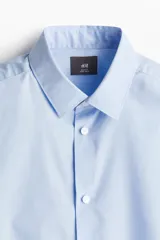 Camisa de tela azul claro con tratamiento de planchado fácil, cuello inglés, tapeta clásica y canesú en la espalda. Mangas largas con tapeta y botón, y cierre ajustable de botón en los puños. Bajo levemente redondeado. Corte ajustado que realza el contorno del cuerpo.