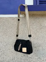 Cartera bandolera de nylon color negro con detalles en charol. Presenta un diseño fruncido con cordones decorativos combinados, grifa de marca en material sintético y correa regulable e intercambiable con logo estampado.