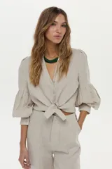 Camisa de lino color beige, con escote en V, cuello mao y mangas abullonadas tres cuartos. Cuenta con cierre frontal de botones y tiras ajustables para atar en la cintura.
