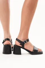 Sandalia de cuero negro con textura trenzada, taco ancho, plataforma baja y tira con hebilla ajustable al tobillo. Presenta pequeños detalles de tachas metálicas en la tira delantera.