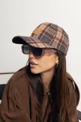 Gorra de estilo béisbol con estampado de cuadros tipo tartán en tonos marrones y grises. Presenta un parche frontal de cuero sintético con la marca grabada y cierre ajustable en la parte posterior.