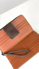 Billetera grande tipo sobre con correa de mano, confeccionada en cuero marrón con detalle central de cuero con pelo vacuno en tonos marrón, negro y blanco. Presenta cierre perimetral y solapa frontal con broche magnético y logo de búho grabado.