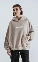 Hoodie oversize de algodón color beige, con capucha forrada, bolsillo canguro frontal y estampado con la palabra AMOR en el pecho. Presenta costuras expuestas en las mangas y un acabado deshilachado en el ruedo.