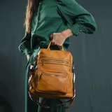 Mochila de símil cuero color marrón con apertura superior y tres bolsillos frontales con cierres metálicos. Cuenta con bolsillos laterales y una correa de galón con diseño geométrico, regulable y extraíble, que permite su uso como bandolera.
