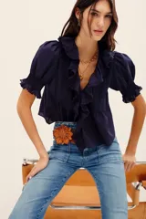 Camisa azul marino de algodón, con mangas cortas abullonadas y puños fruncidos con elástico. Presenta un amplio escote en V adornado con volados con detalles bordados y ojales a lo largo de la botonera frontal.